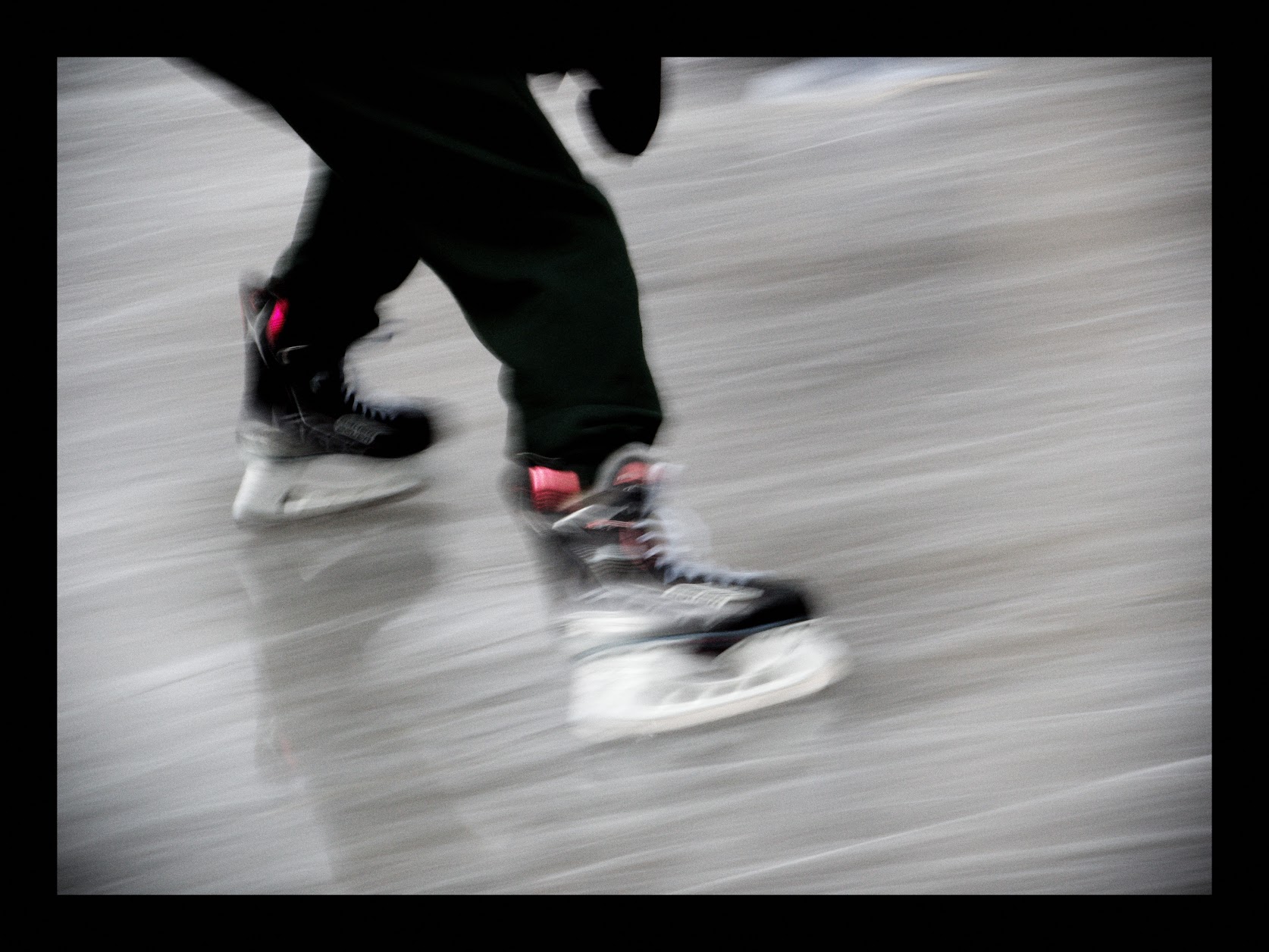 A low-angle photograph of a skater's legs crossing an ice rink, panned so that both the skater and the ice are smeared into horizontal streaks. Black skates with a flash of pink at the ankle, dark trousers, and a pale grey rink; almost nothing in the frame is in focus. Black border.