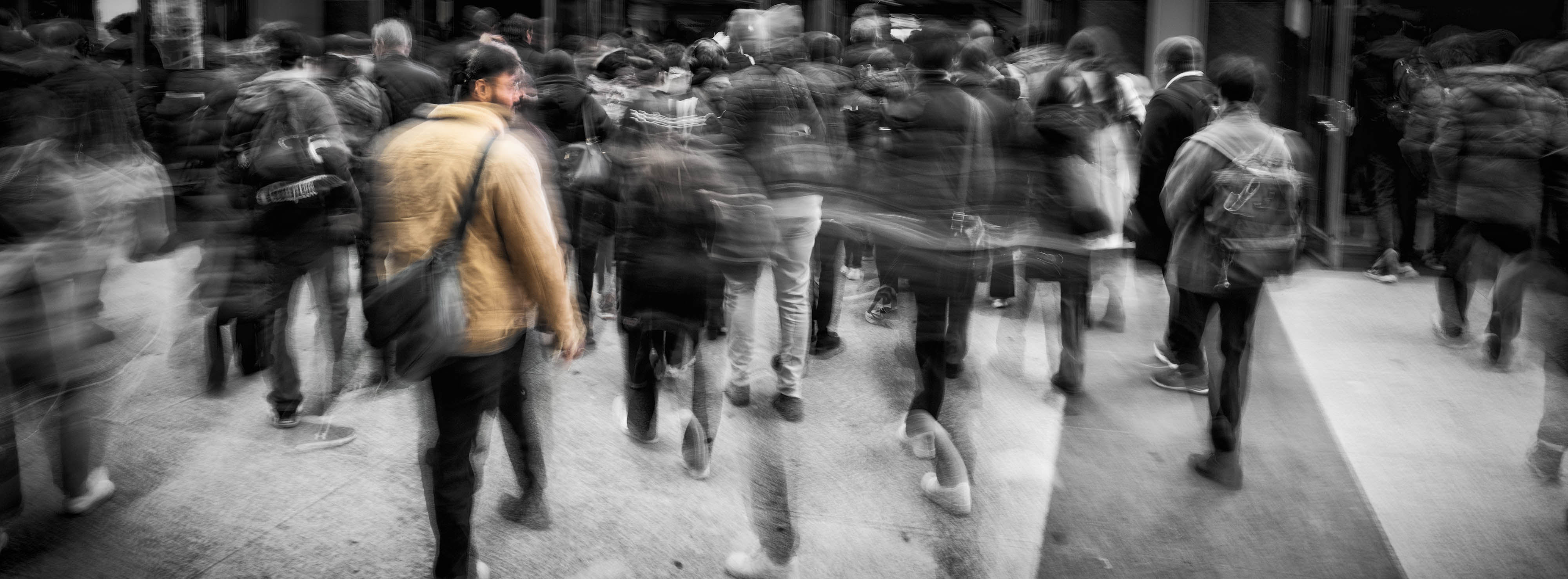 A very wide panoramic photograph of a crowd of pedestrians crossing a pavement, shot with a long exposure so most of the figures blur into overlapping ghosts. The image is black and white apart from one woman near the left in a mustard yellow jacket, who is the only colour in the frame and the point where the eye comes to rest.