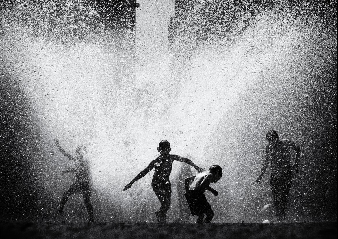A high-contrast black and white photograph of four children playing in the huge spray of a burst fountain or open hydrant. The water erupts upward and outward in a bright cloud of droplets that fills most of the frame, backlit so the children read as dark silhouettes against it. One child in the centre runs with both arms flung wide, another ducks away beside them, a third throws up an arm on the left, and a fourth stands further to the right; a tall building looms faintly through the spray behind them.