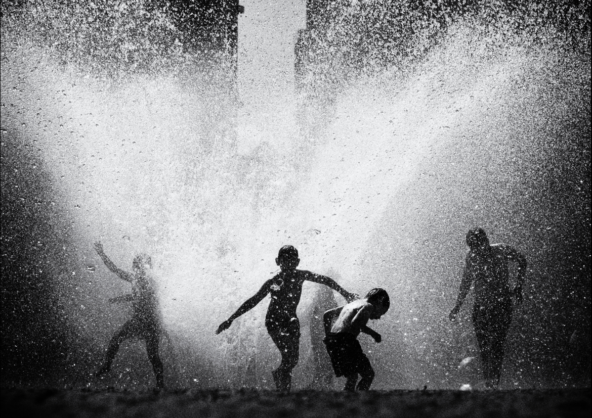 A high-contrast black and white photograph of four children playing in the huge spray of a burst fountain or open hydrant. The water erupts upward and outward in a bright cloud of droplets that fills most of the frame, backlit so the children read as dark silhouettes against it. One child in the centre runs with both arms flung wide, another ducks away beside them, a third throws up an arm on the left, and a fourth stands further to the right; a tall building looms faintly through the spray behind them.