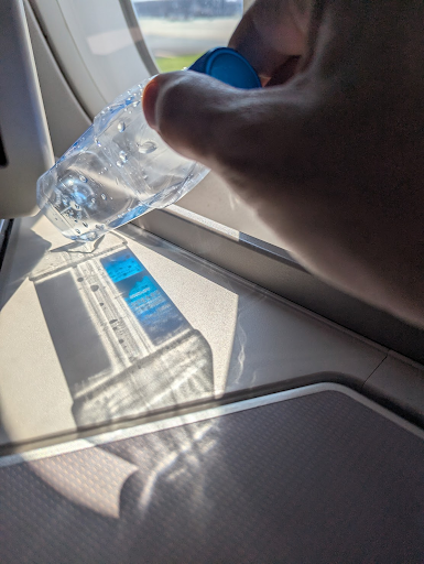 A hand holds a crumpled, half-empty clear plastic water bottle with a blue cap up against an aeroplane window. Bright low sunlight passes through the bottle and throws two things onto the pale window ledge below: a hard dark shadow and a smear of refracted, faintly coloured light. Green ground is just visible through the window behind the hand.