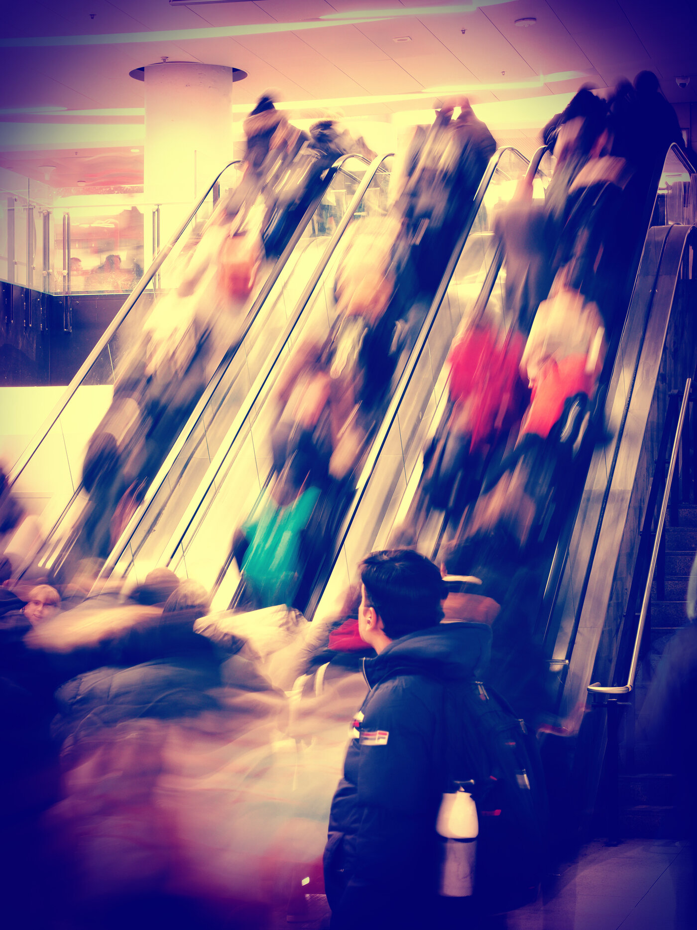 A bank of crowded escalators in a mall or transit hall, the moving people blurred into vertical streaks and the whole image toned warm magenta and gold. At the bottom centre a young man in a dark jacket stands almost sharp and still, looking off to one side - the one fixed point in all the motion.