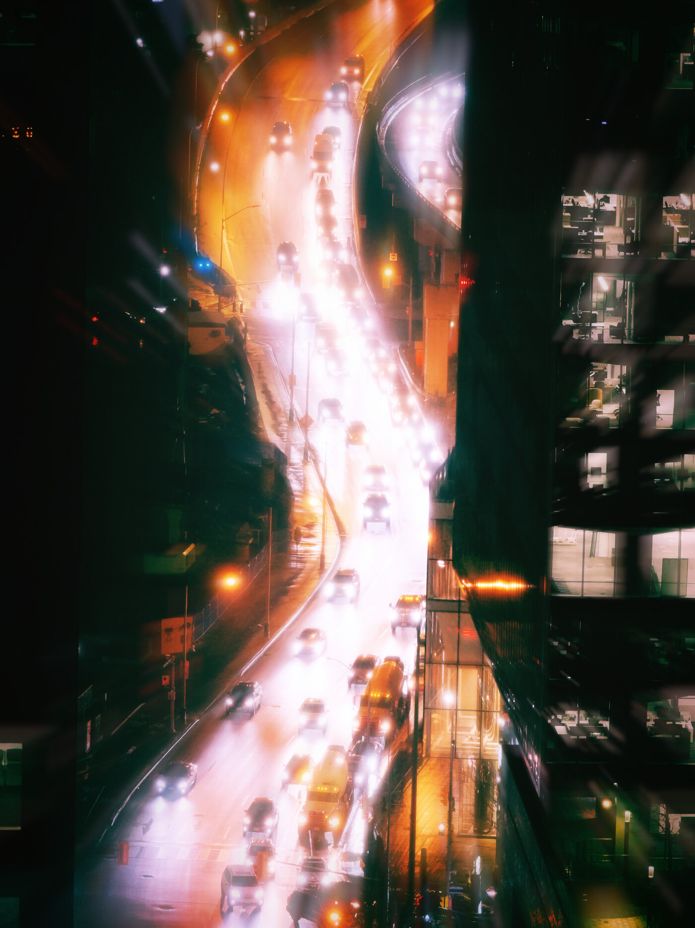 A curving city expressway at night seen from high above, framed in a narrow vertical slot between two dark tower edges, lit office windows stacked down the right-hand side. The lanes of traffic blur into a bright river of white and amber light, its core blown almost to pure white where the road bends away.