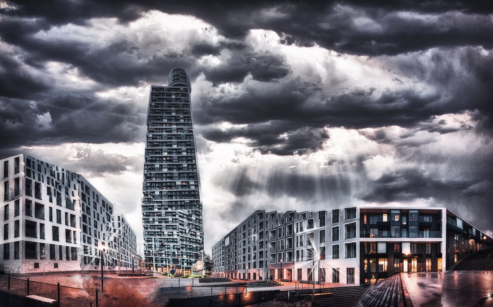 A wide view of a slim high-rise tower in heavy grey cloud, flanked by lower apartment blocks whose facades lean inward, above a wet plaza reflecting streetlight sunstars. The scene looks like a single dramatic photograph but is assembled from many frames shot from different positions.