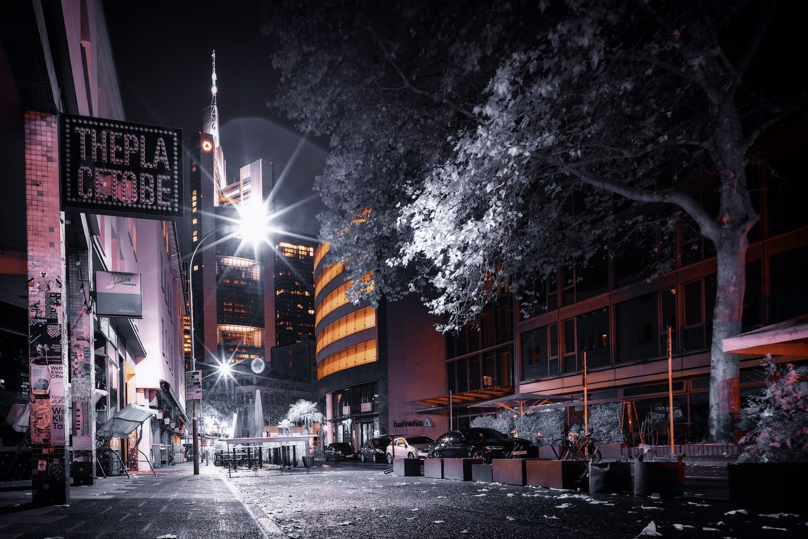 A night street in Frankfurt shot at extreme wide angle in infrared. A lit marquee sign reading THE PLACE TO BE hangs on the left, the Commerzbank tower's spire rises in the centre distance, and a large tree on the right has gone silver-white the way foliage does in infrared. Streetlights flare into sharp sunstars and the whole scene sits in pink and sodium orange.