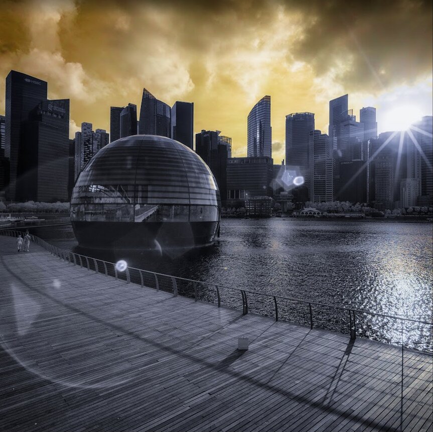 Infrared shot taken in Singapore. Sky is yellow, much of the picture is grey-blue. There is a skyline in the back, a bay in the middle and a wooden walkway in the front. At the left middle there's a sphere in the water (it houses Singapore's flagship Apple Store). There's a lens flare and a sun star.